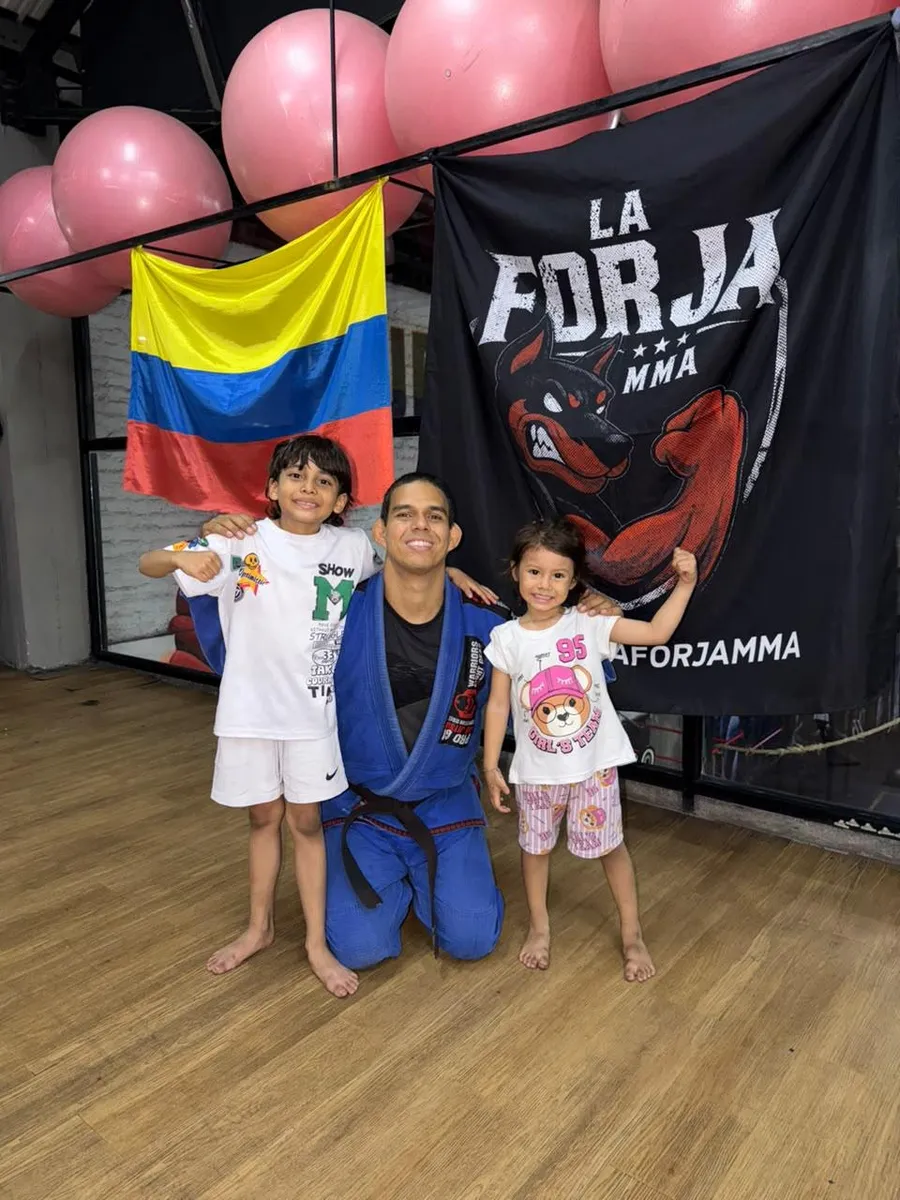 Coach de BJJ con dos alumnos de BJJ Kids frente a la bandera de La Forja MMA, Cali
